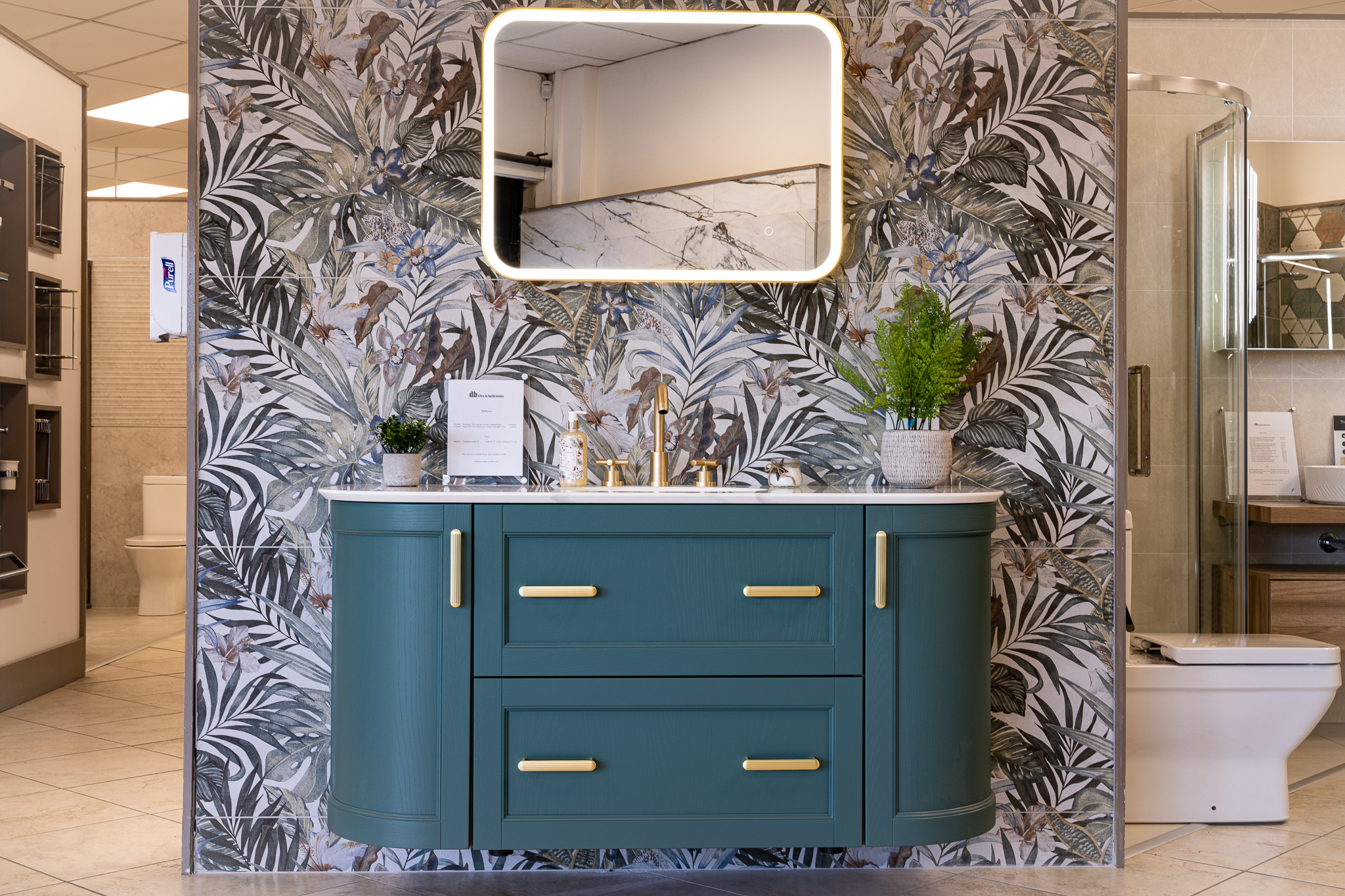 A curved teal vanity unit with brass handles and tap, set against tropical leaf-pattern wall tiles under an illuminated mirror