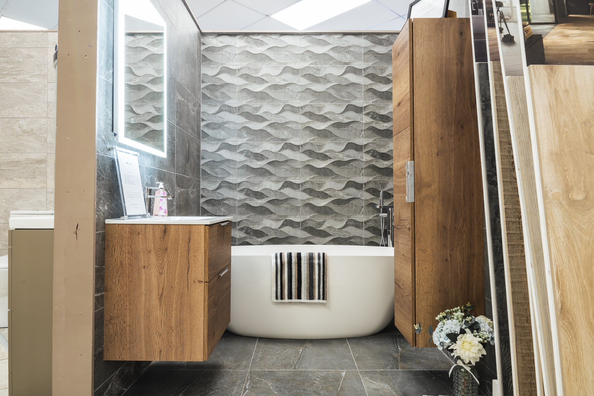 A freestanding bath in front of a grey wave-pattern feature wall, with an oak-effect vanity and tall unit on dark stone-effect floor tiles