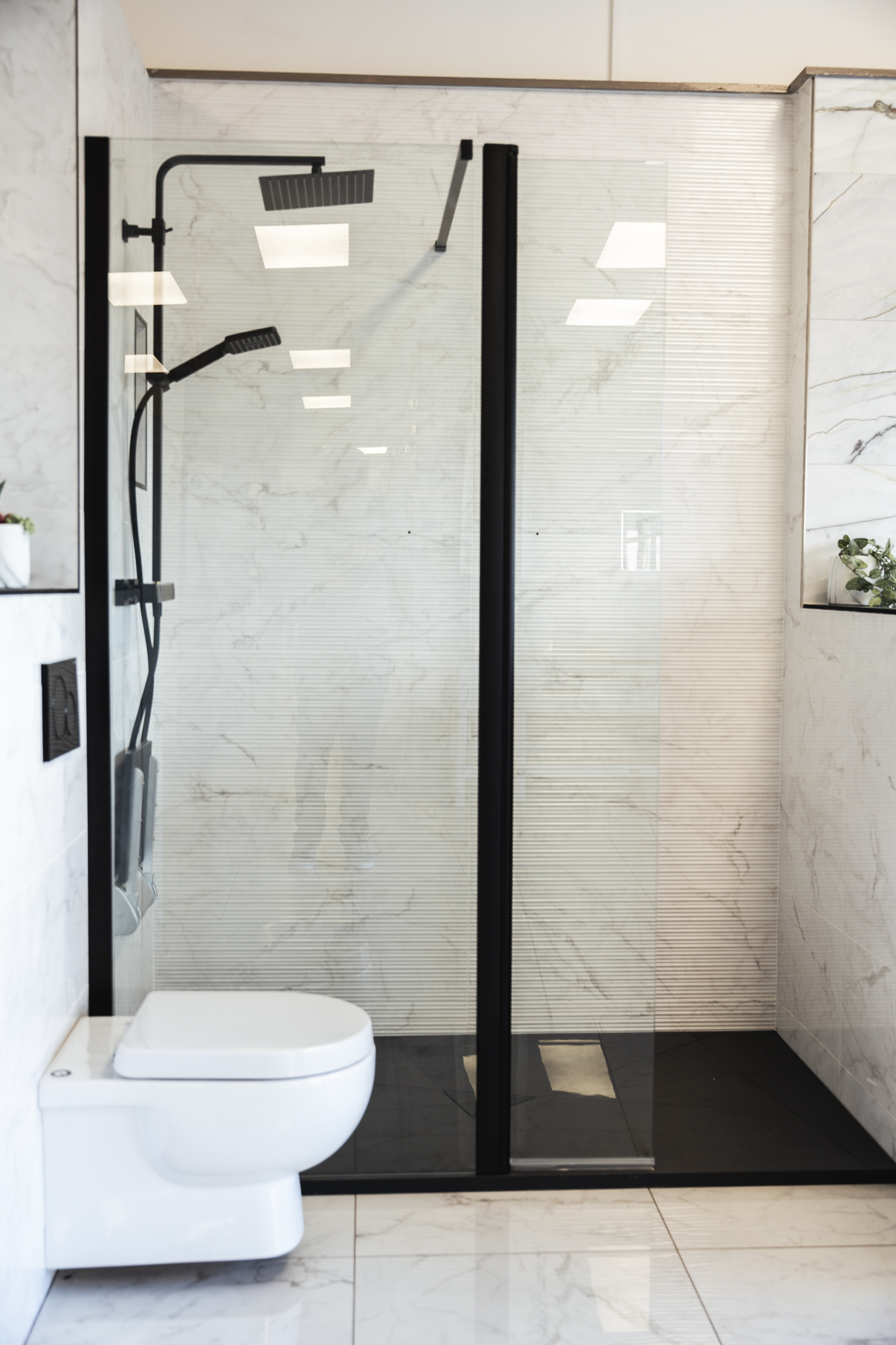 A walk-in shower with a black frame and black shower tray beside a wall-hung WC, with marble-effect tiles on the walls and floor